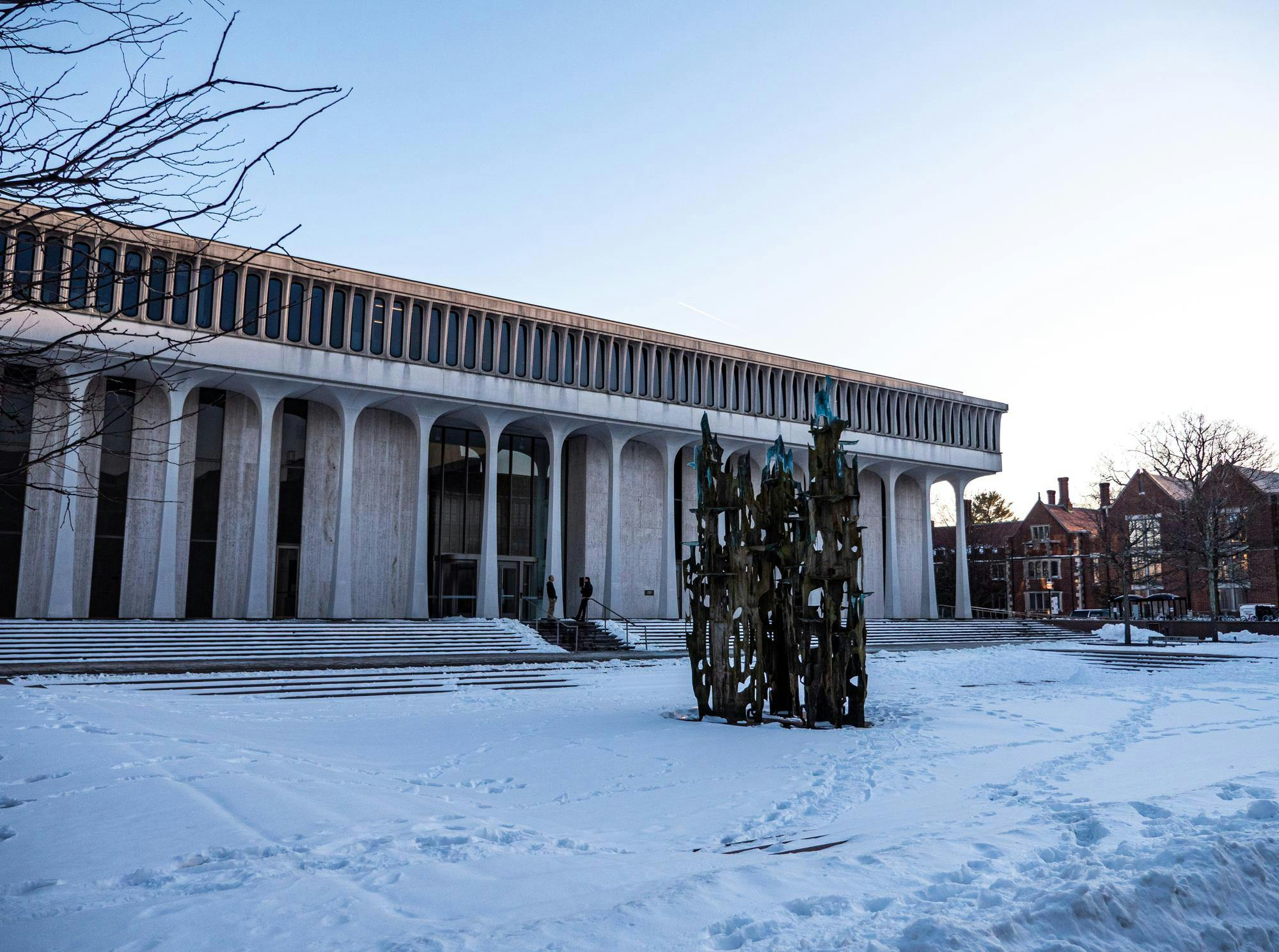 A large building with white columns stands behind a gray structure. The ground is covered in snow.