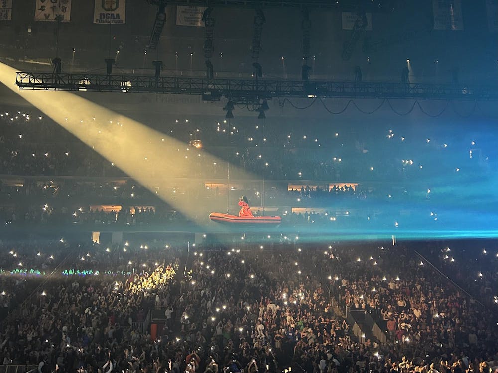 A woman in a red garment stands on a platform, suspended over a large crowd of people holding up flashlights.