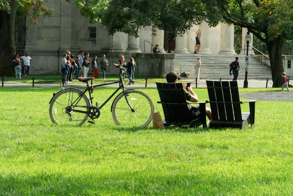 A wide shot of a lawn with a bicycle and two Adirondack chairs in the foreground. A group of students mill in the background in front of a white marble building. 