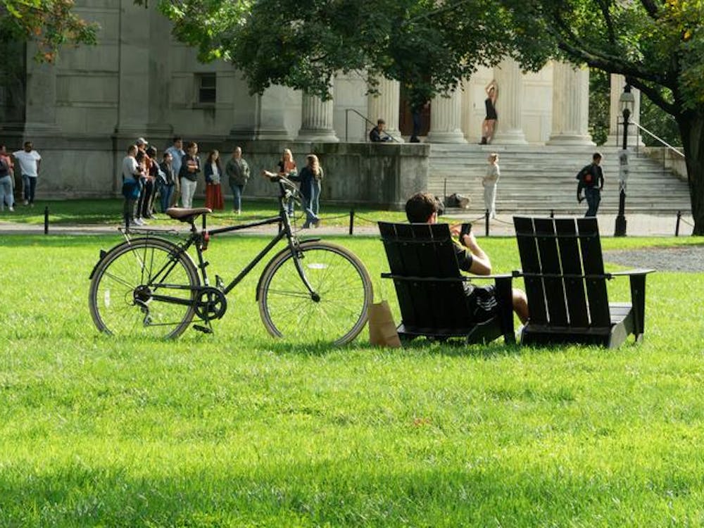 A wide shot of a lawn with a bicycle and two Adirondack chairs in the foreground. A group of students mill in the background in front of a white marble building. 