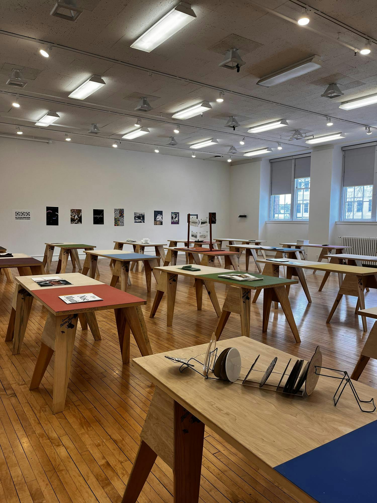 In a room with white walls and windows, rows of colorful desks with books sit. On the rear wall, there is a line of posters. 