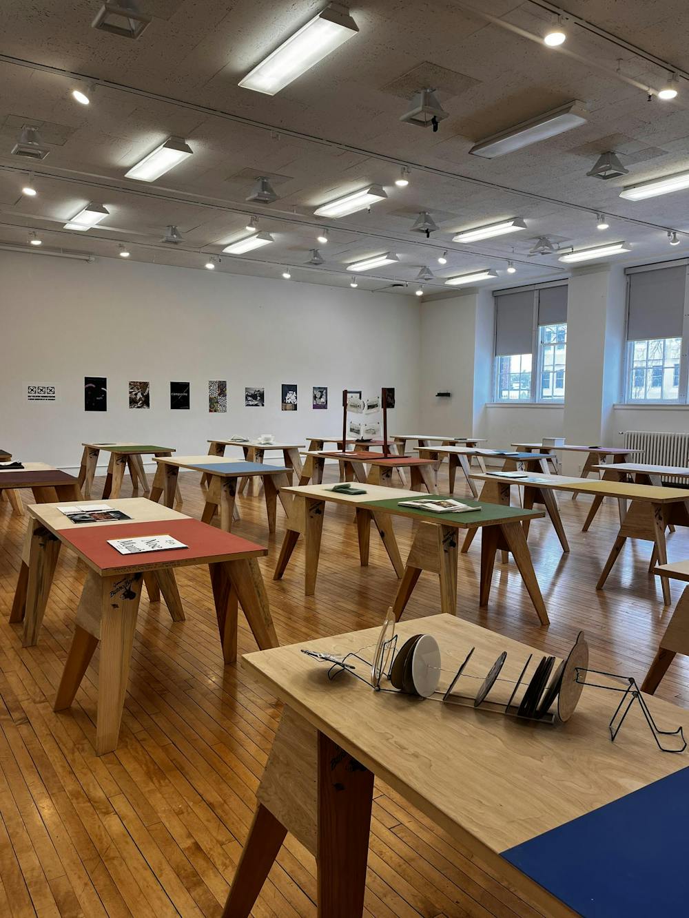 In a room with white walls and windows, rows of colorful desks with books sit. On the rear wall, there is a line of posters. 
