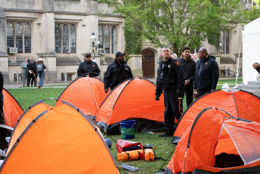 Multiple orange tents stand on the green grass. The tents are in front of a tan building with windows and green trees.