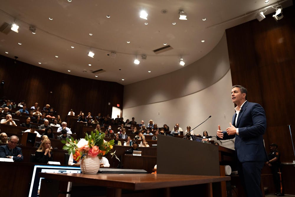 A man in a navy blue suit speaks to a room of people in terraced rows. 