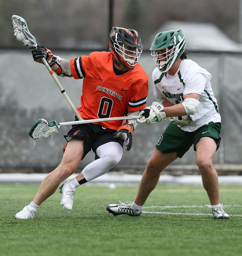 A man in an orange jersey running with a lacrosse stick in hand while being defended by another man in green and white during a lacrosse game.