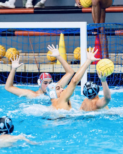 Water polo player shoots a contested shot in the pool with two defenders in front of him.