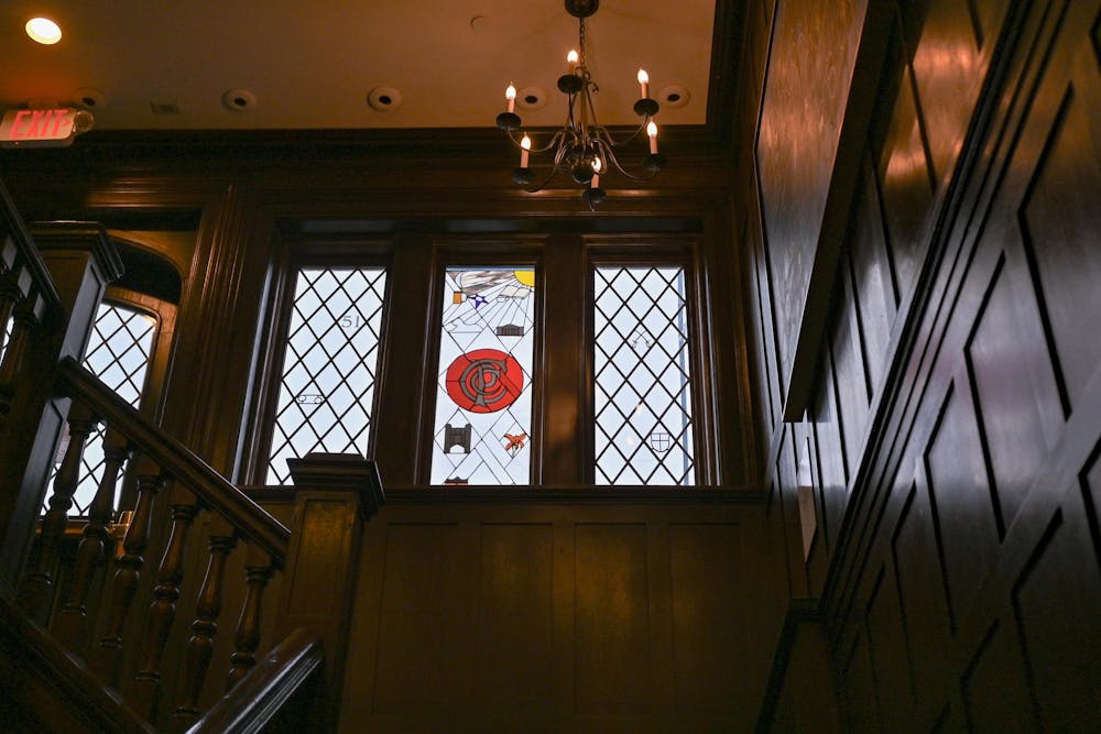 Featured are wooden walls and a wooden staircase. A chandelier is hanging from the sky. In the middle of the wooden walls, there is a stain glass window with a “C” symbol for “Campus Club.”