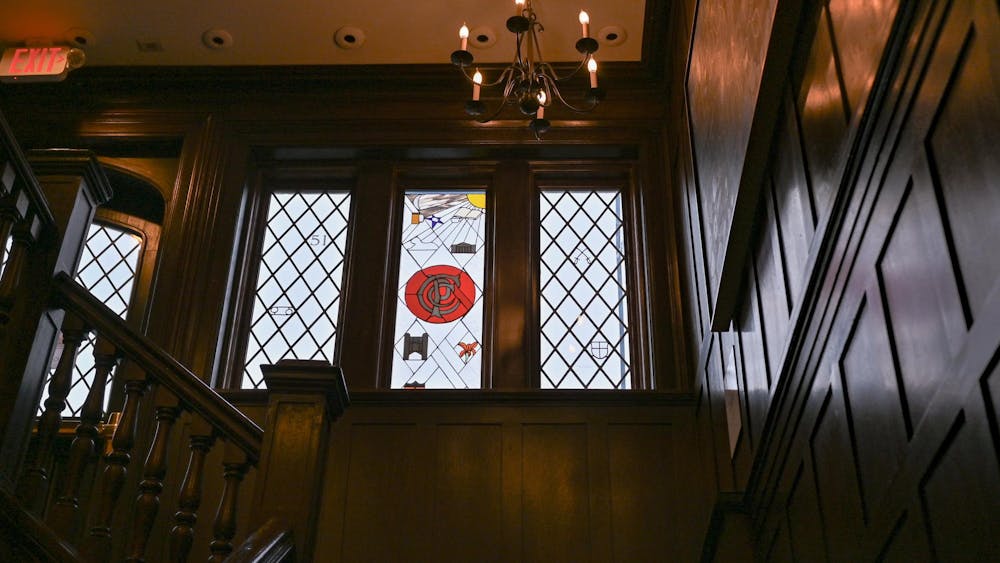 Featured are wooden walls and a wooden staircase. A chandelier is hanging from the sky. In the middle of the wooden walls, there is a stain glass window with a “C” symbol for “Campus Club.”