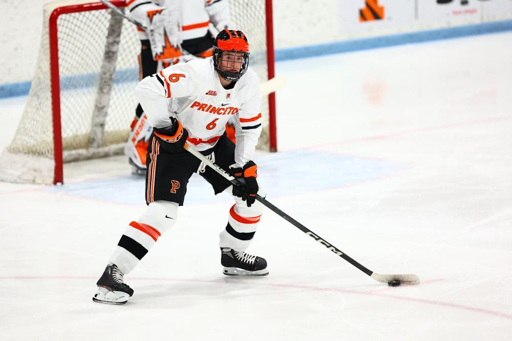 Man in orange and white uniform holds puck in defensive zone, looking up ice to teammates. 