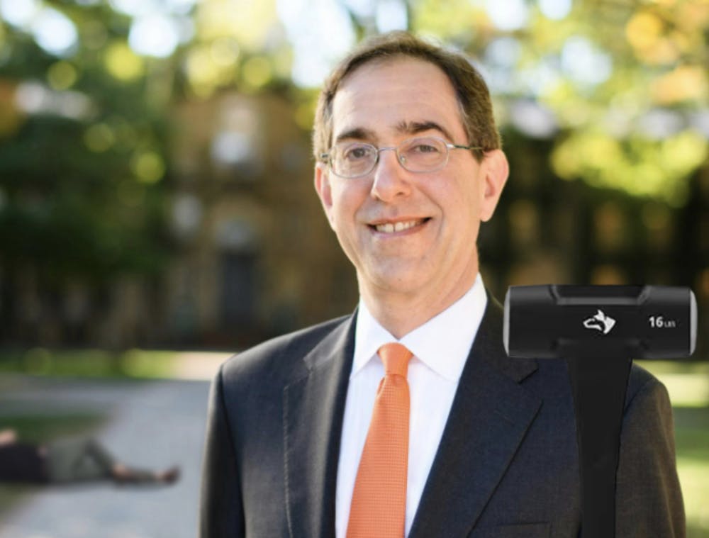 Portrait photo of Princeton University President Christopher Eisgruber standing in front of Nassau Hall, holding a sledgehammer with a immobilized student lying in the background.