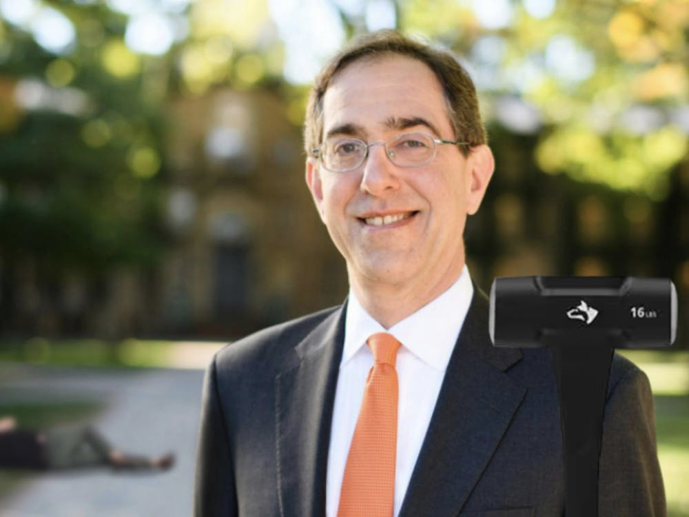 Portrait photo of Princeton University President Christopher Eisgruber standing in front of Nassau Hall, holding a sledgehammer with a immobilized student lying in the background.