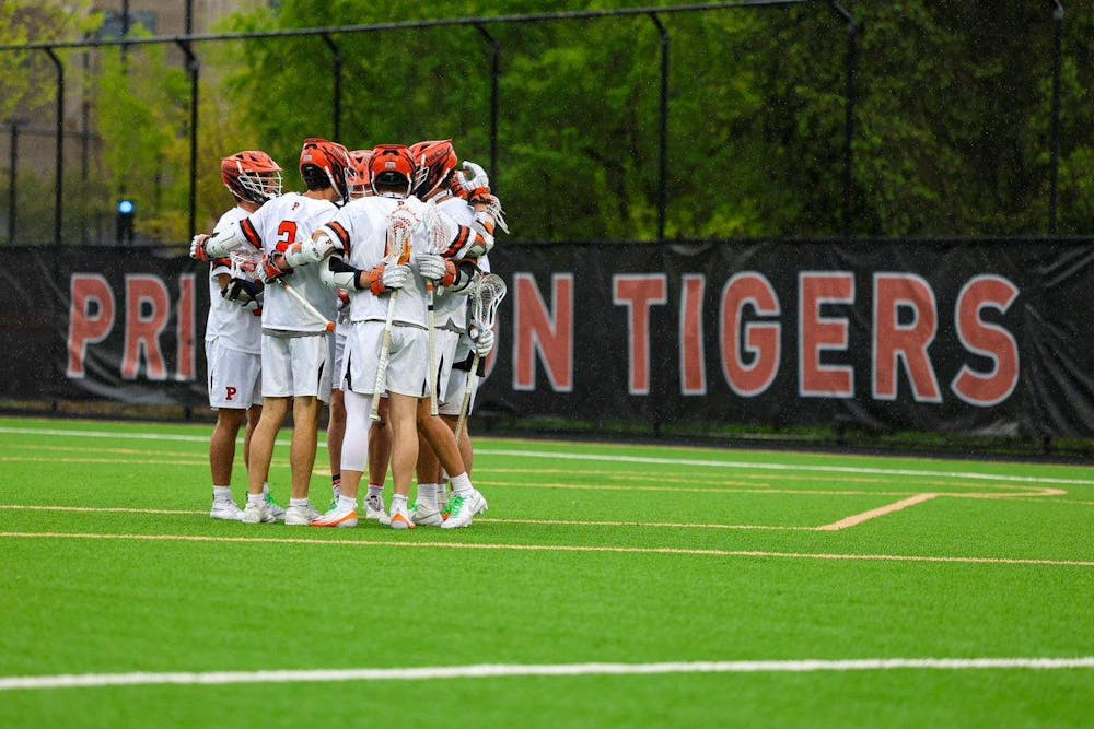 A group of men’s lacrosse players in white uniforms and orange helmets huddle together on a grass field.