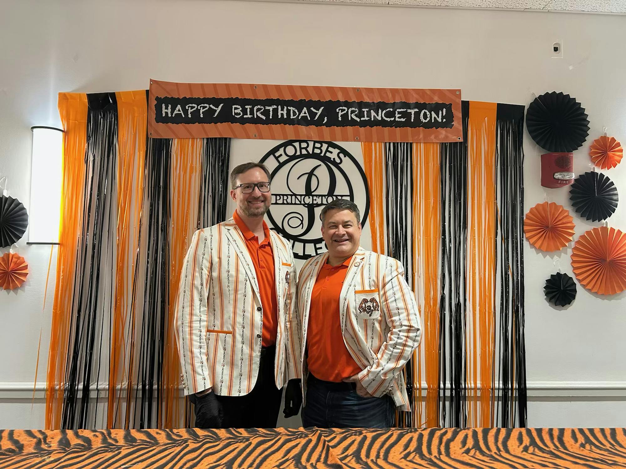 Two alumni dressed in orange and black stand in front of a sign saying "Happy Birthday, Princeton!"