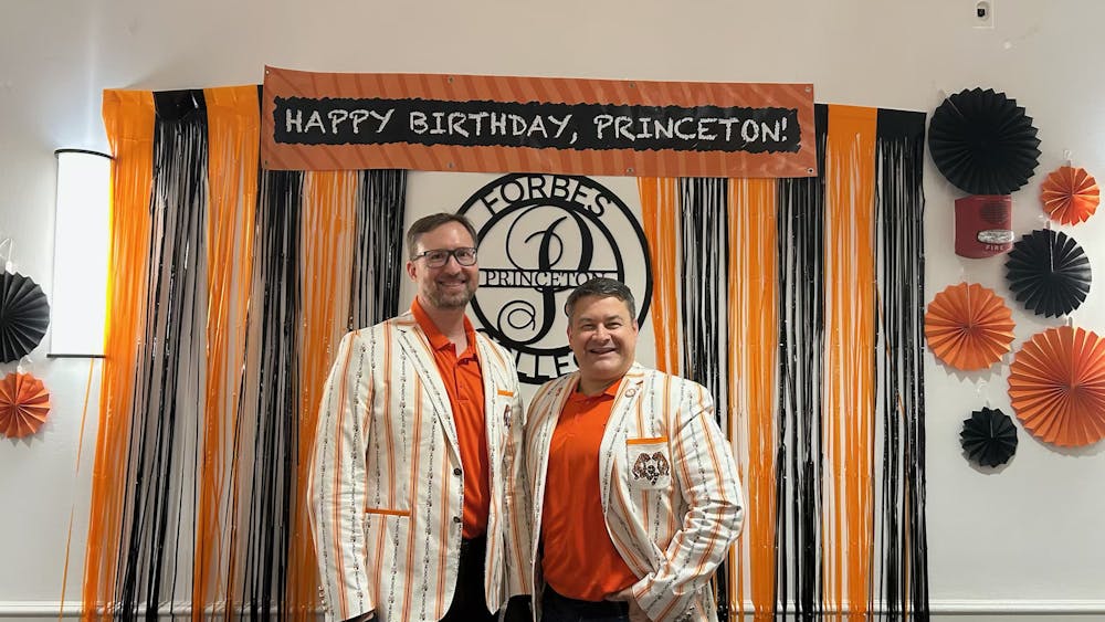 Two alumni dressed in orange and black stand in front of a sign saying "Happy Birthday, Princeton!"