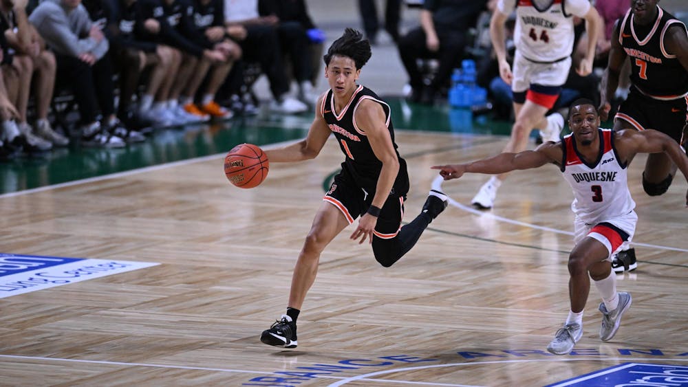 Male basketball player in an orange and black jersey runs down the court with a basketball, pursued by the other team.