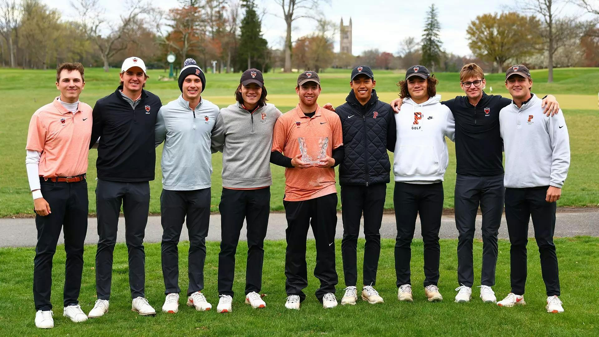A group of golfers stand arm in arm smiling on golf course with the golfer in the center holding a transparent glass trophy.
