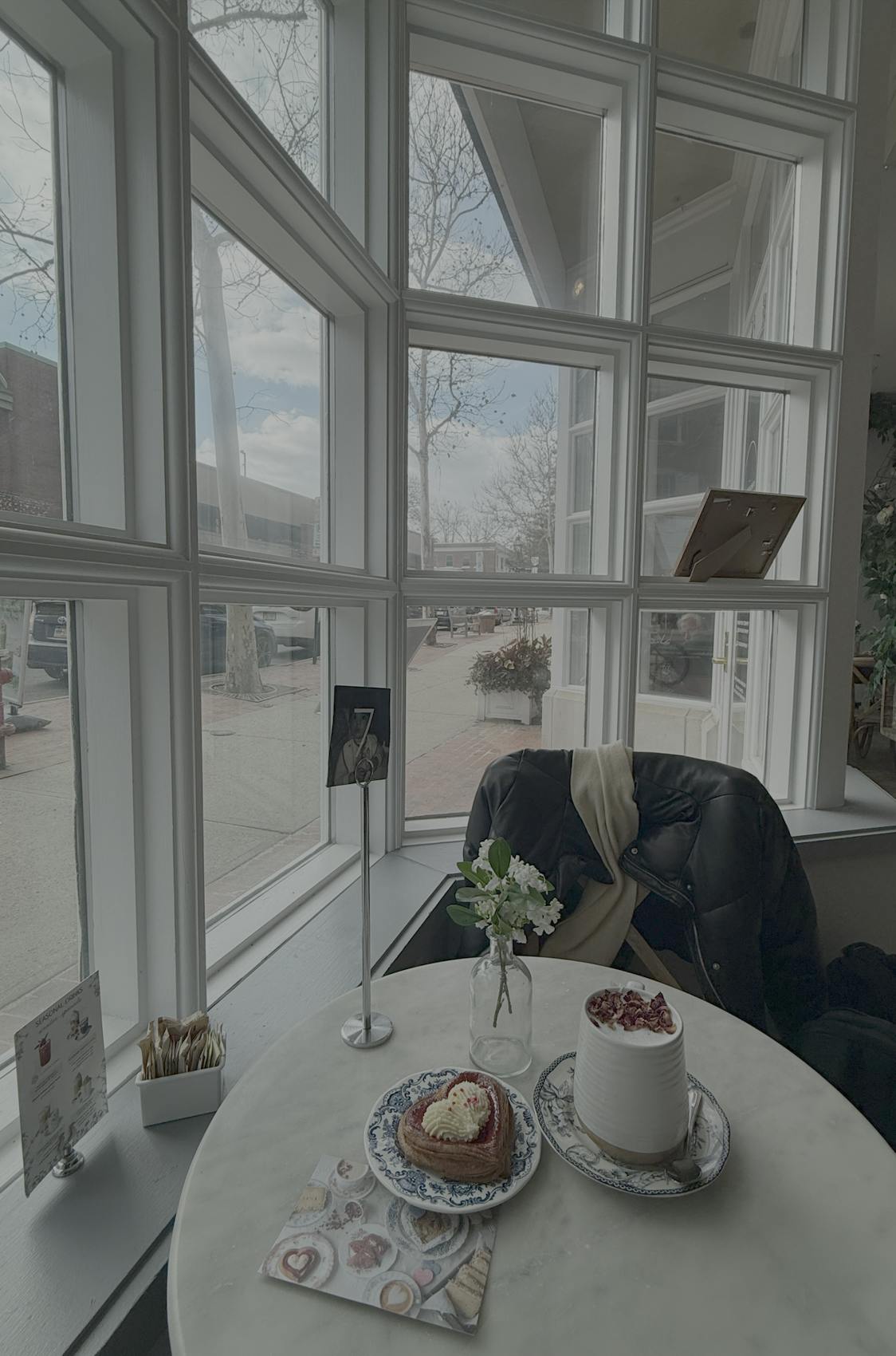 A mug and a pastry sit on a table by a large window with a view into the street. 
