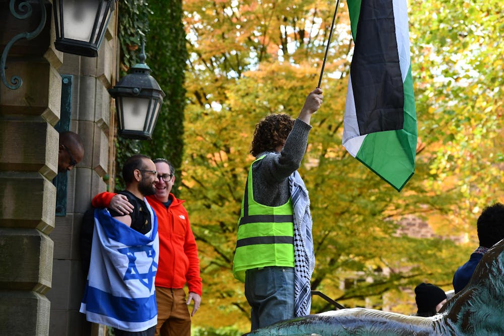 Standing behind a protestor wearing a green work vest, a keffiyeh, and holding a Palestinian flag, a man wraps himself in an Israeli flag on the steps in front of Nassau Hall. 