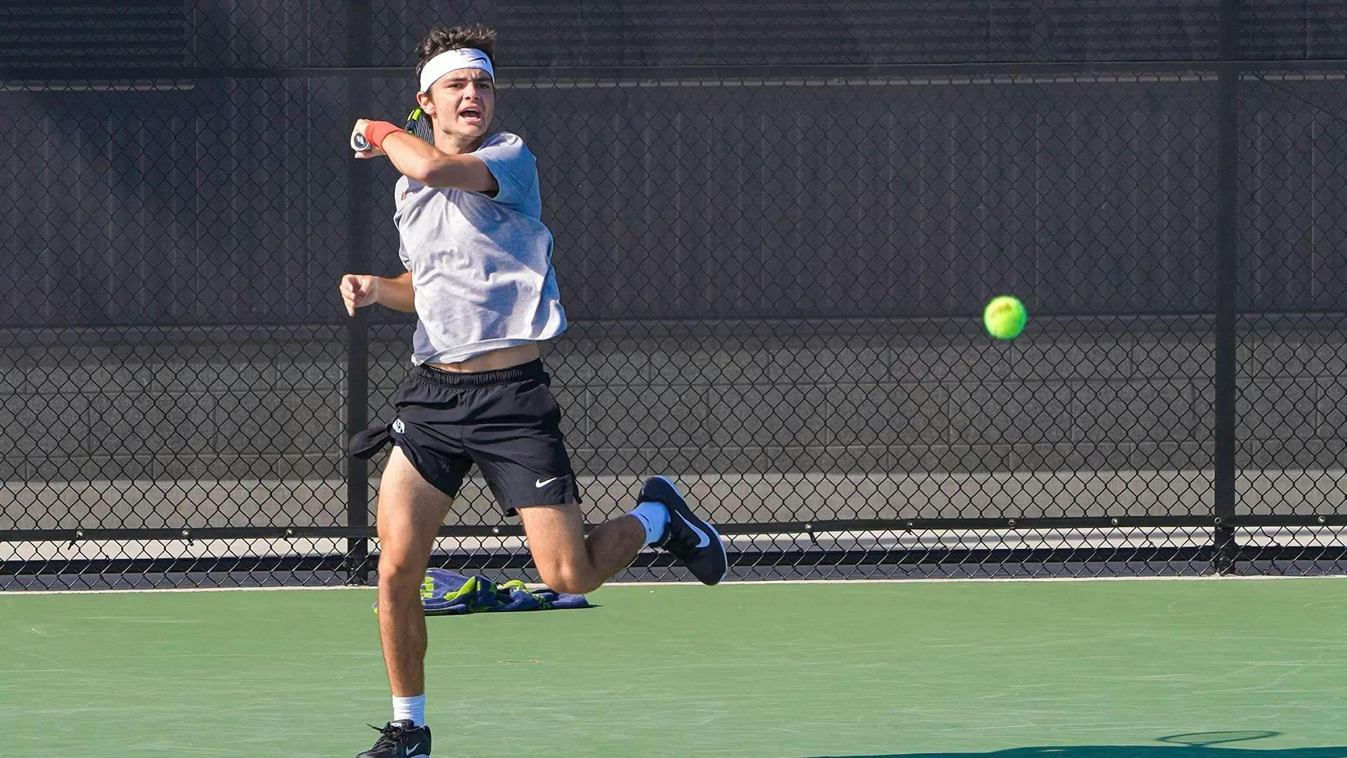 A tennis player in a white shirt and black shorts hits a tennis ball.