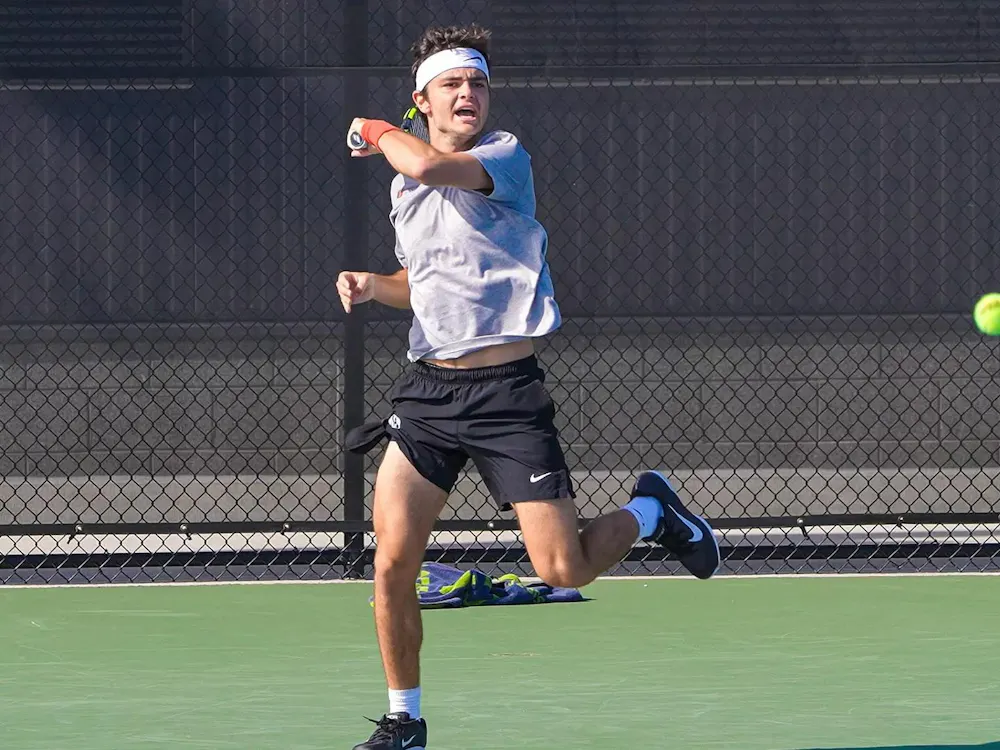 A tennis player in a white shirt and black shorts hits a tennis ball.