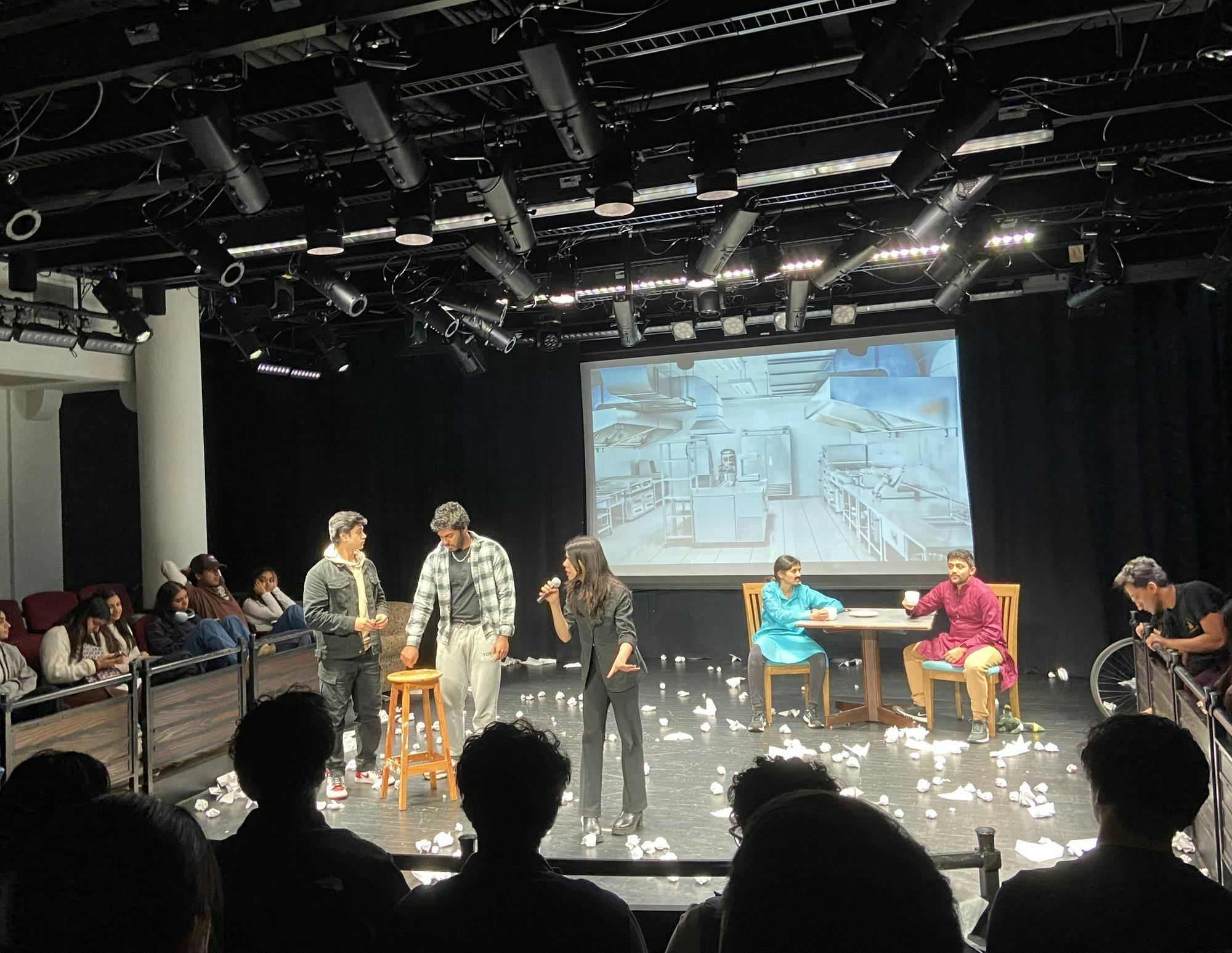 Five people on a stage performing under spotlights. Three people stand around a stool and two other people are sitting at a table.