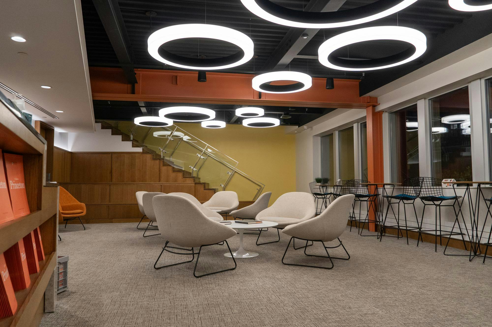 White circle lights over white chairs around low tables on a carpeted floor. There are stairs in the background against a yellow wall.