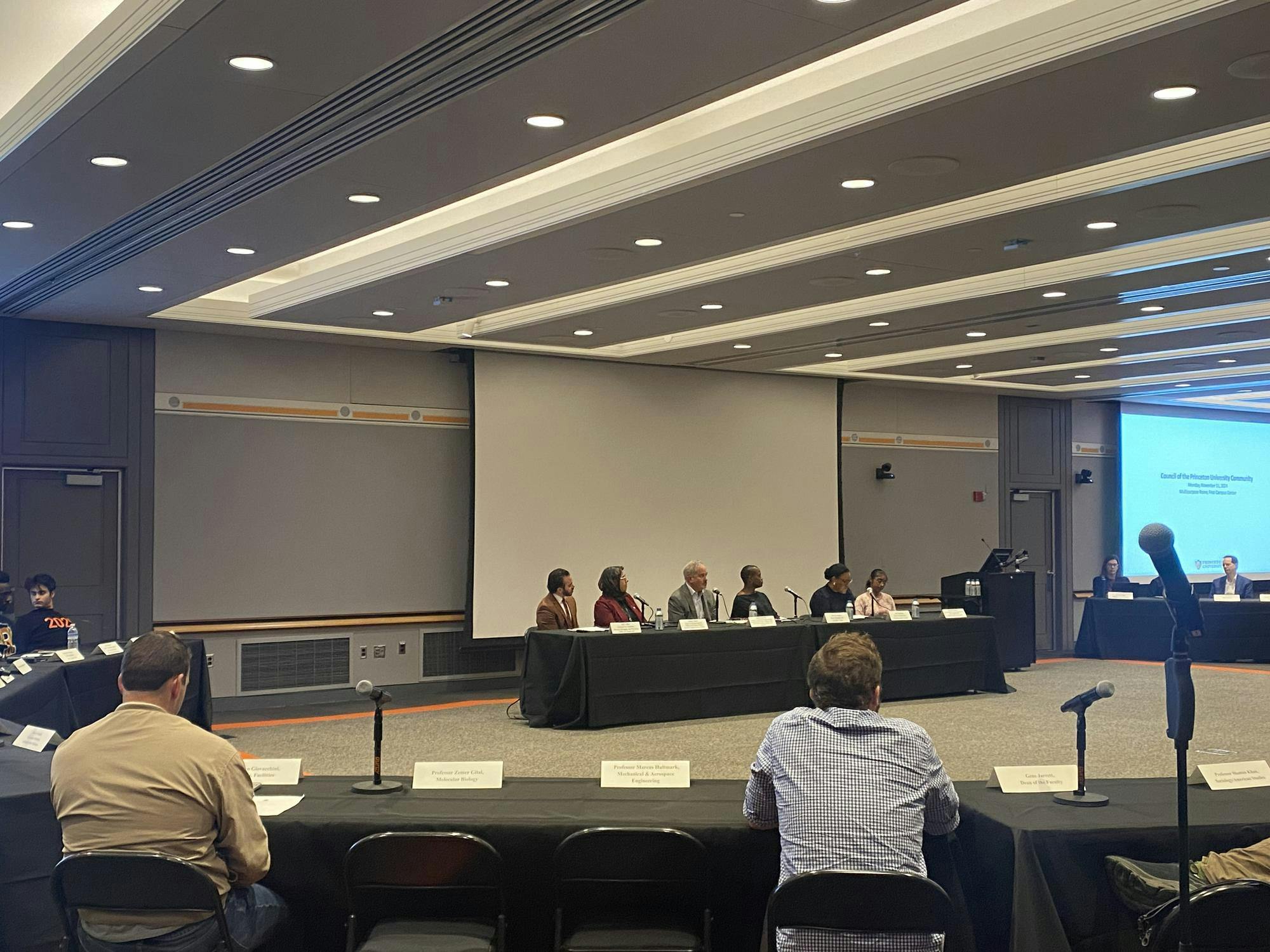 A panel of people at a large meeting room, surrounded by other members of the group at tables.
