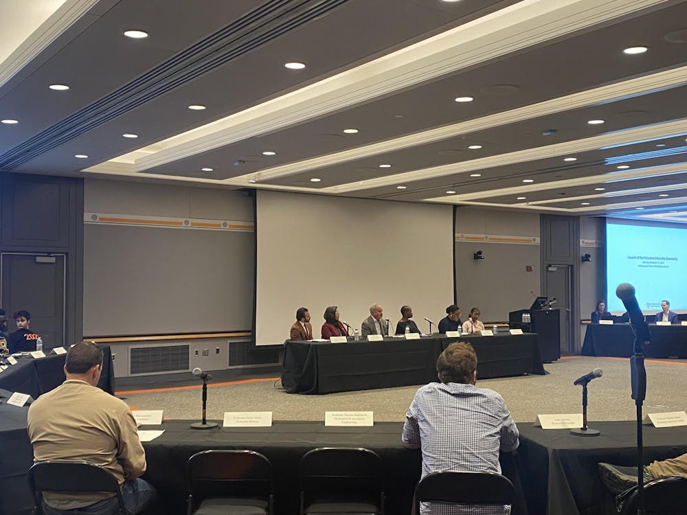 A panel of people at a large meeting room, surrounded by other members of the group at tables.