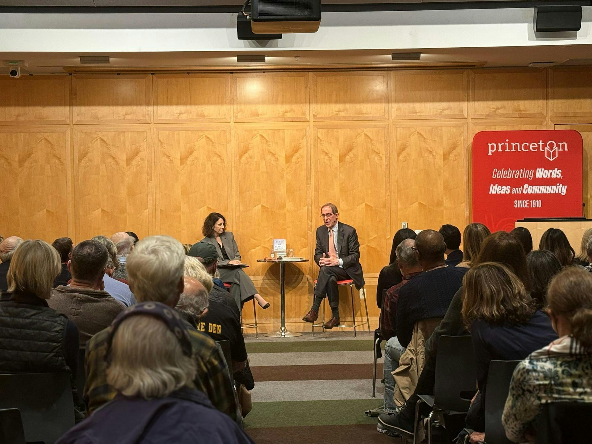 A man and woman are sat at a table. He is talking. There is a crowd of people sitting in the audience listening.