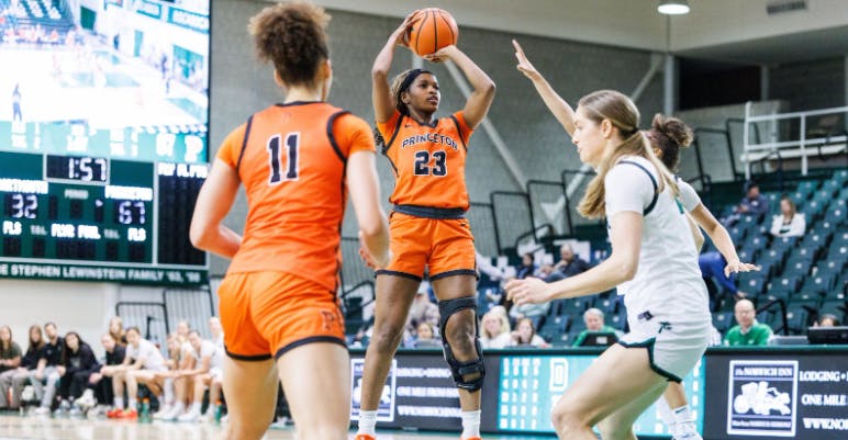 A player wearing an orange jersey is shooting midair over a player in a white jersey.