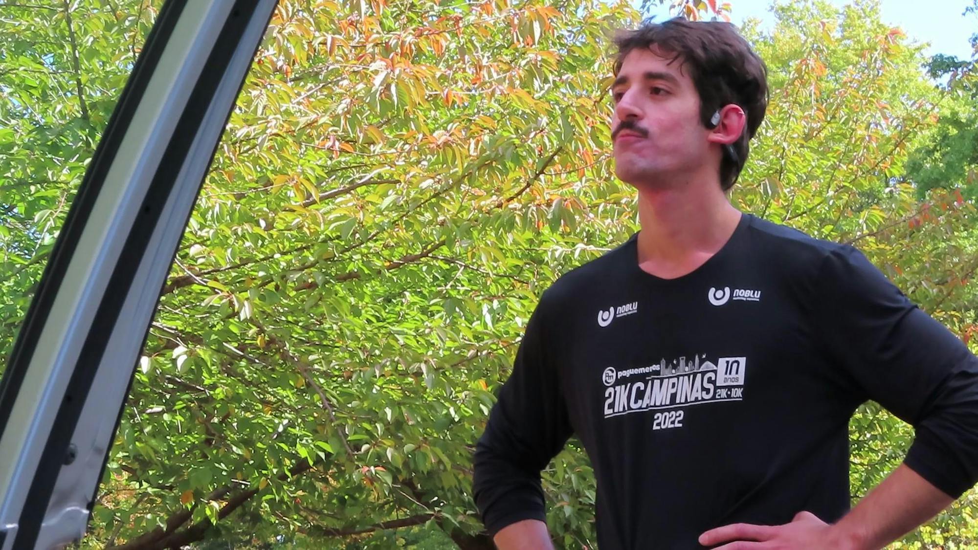 A man wearing a long-sleeved black shirt stands turned slightly the left of the camera, face slightly scrunched, hands on his hips, against a background of green leaves.