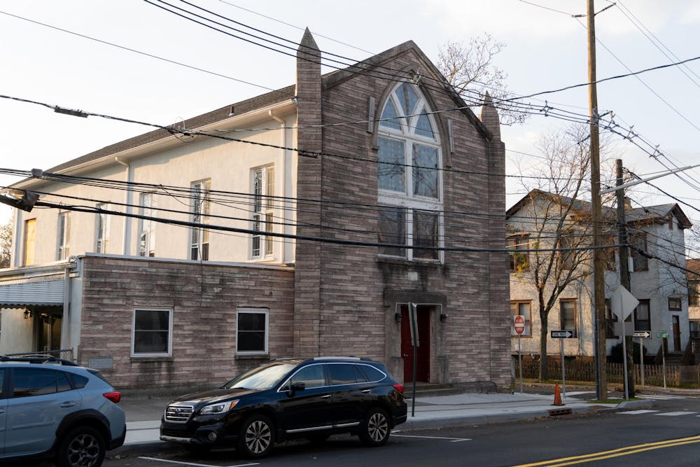 A brown stone church building sits at the corner of Witherspoon and Maclean Streets in Princeton. The sun is setting and there is a black Subaru Outback parked in front.