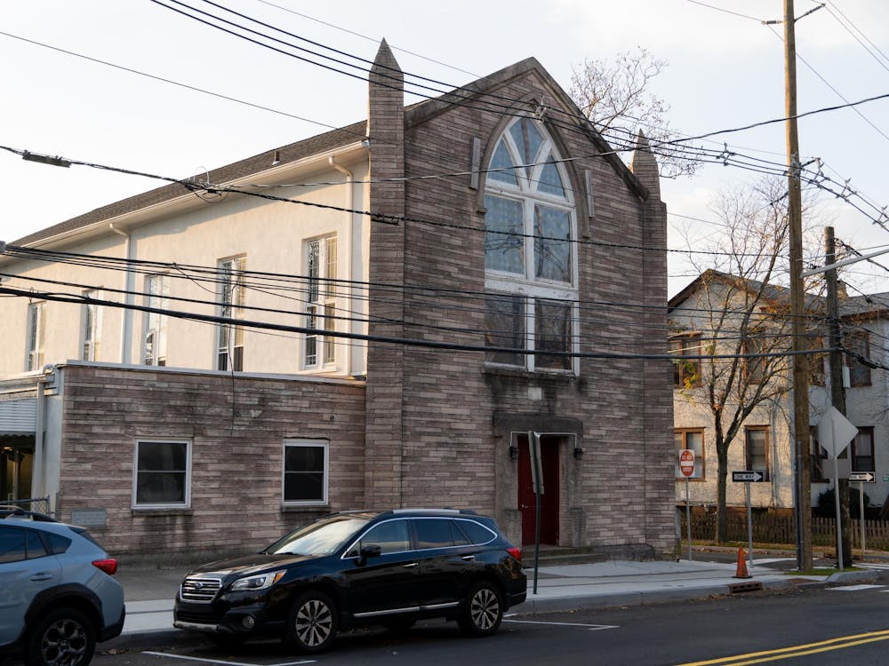 A brown stone church building sits at the corner of Witherspoon and Maclean Streets in Princeton. The sun is setting and there is a black Subaru Outback parked in front.