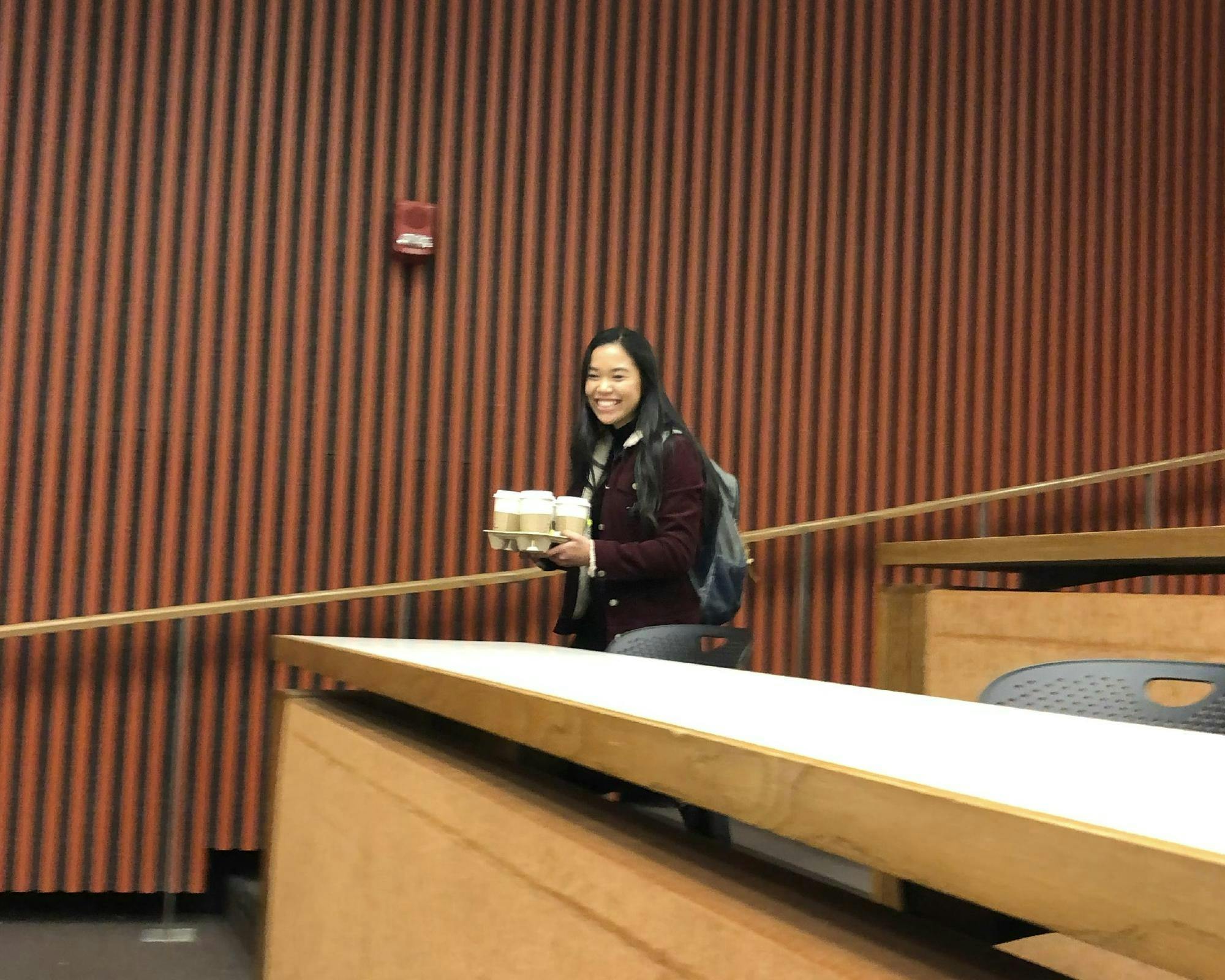 A young woman with a maroon jacket and gray backpack carries a tray of cardboard coffee cups past rows of desks in a lecture hall