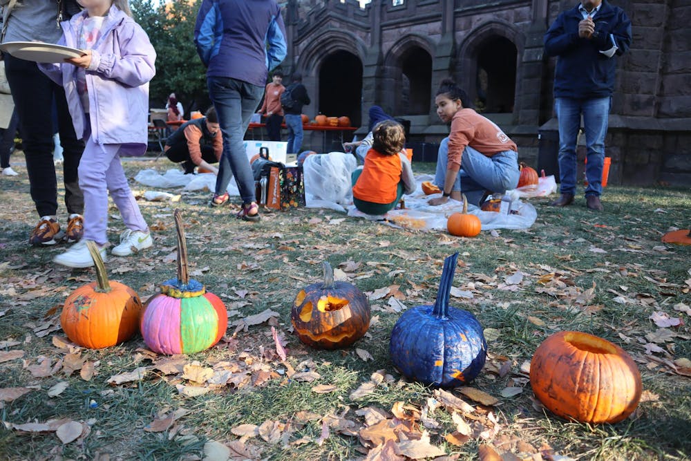 A row of small, brightly painted pumpkins sits in the foreground. In the background, families with children are painting pumpkins.