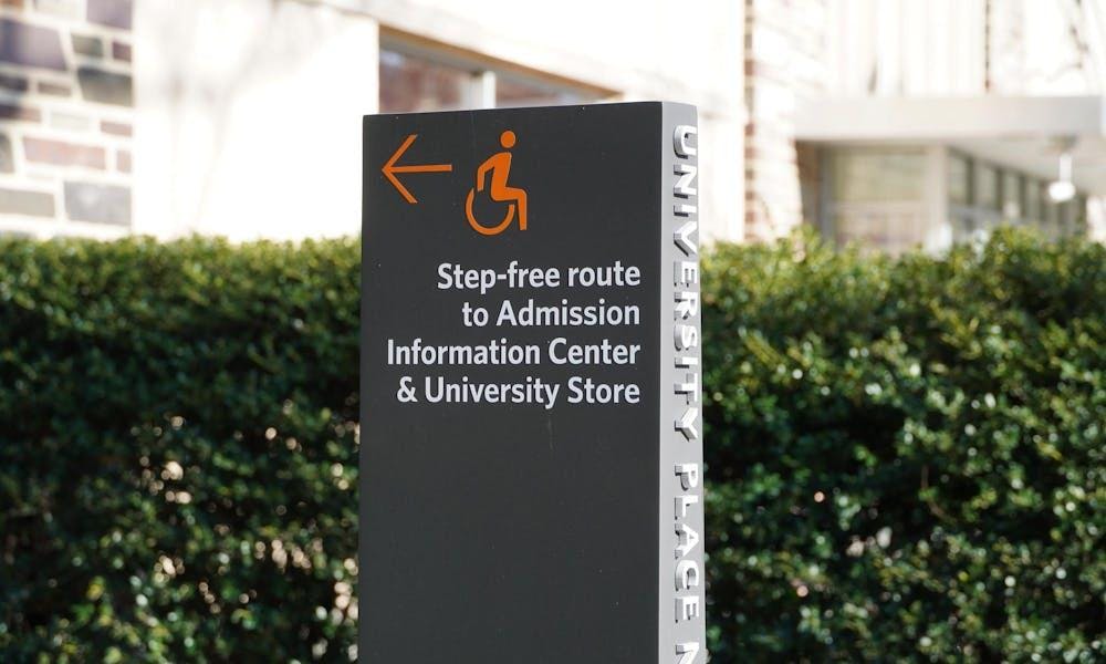 Un cartel gris con escritura blanca delante de un seto verde.