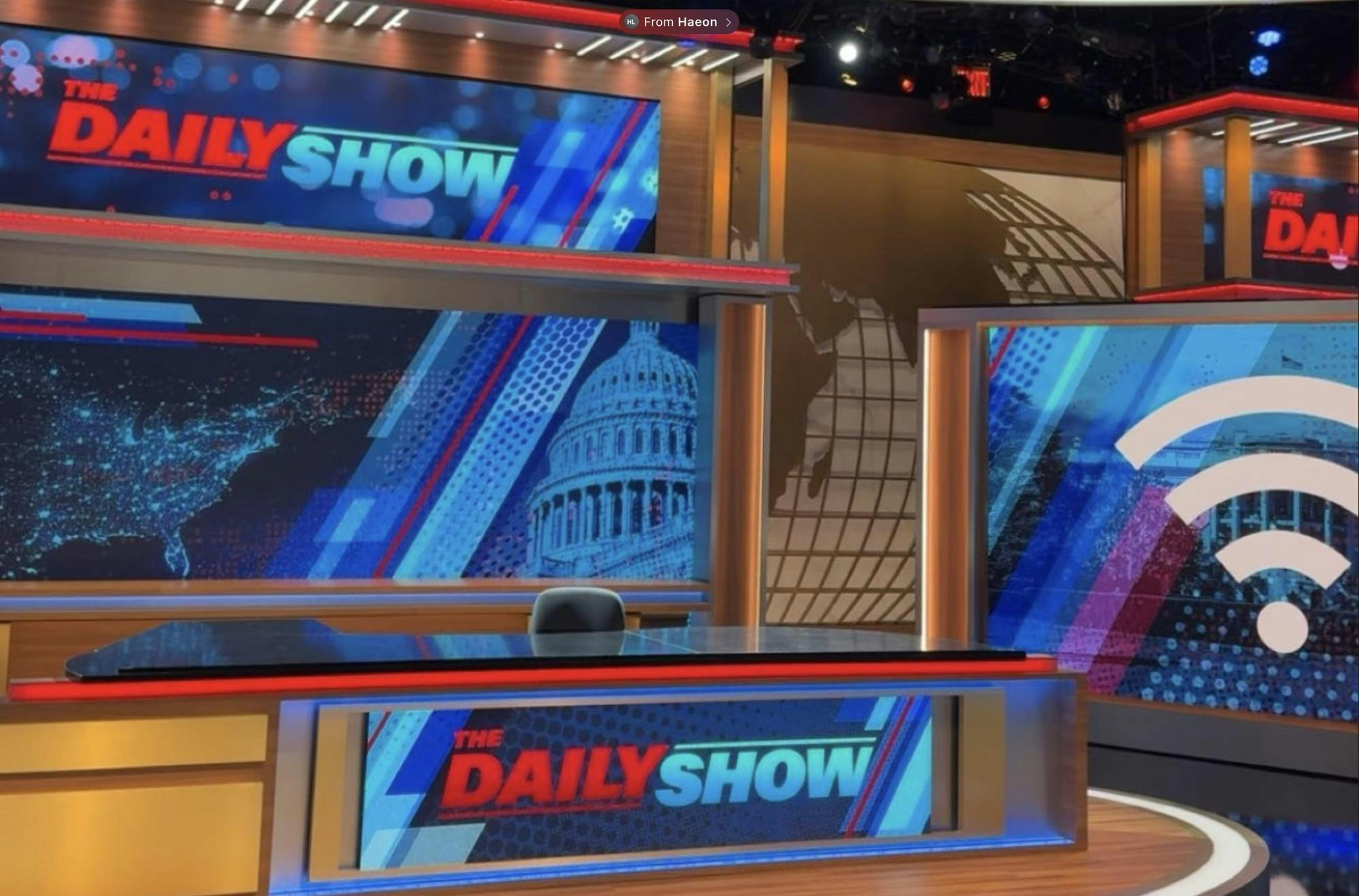 An image of the Daily Show stage featuring the branding of the show on several electronic screens and including a screen in front of the desk.