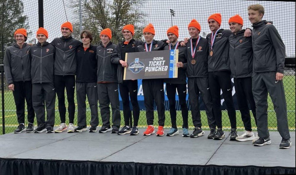 A group of men in black and grey sweatsuits with orange hats stand together holding a sign. 