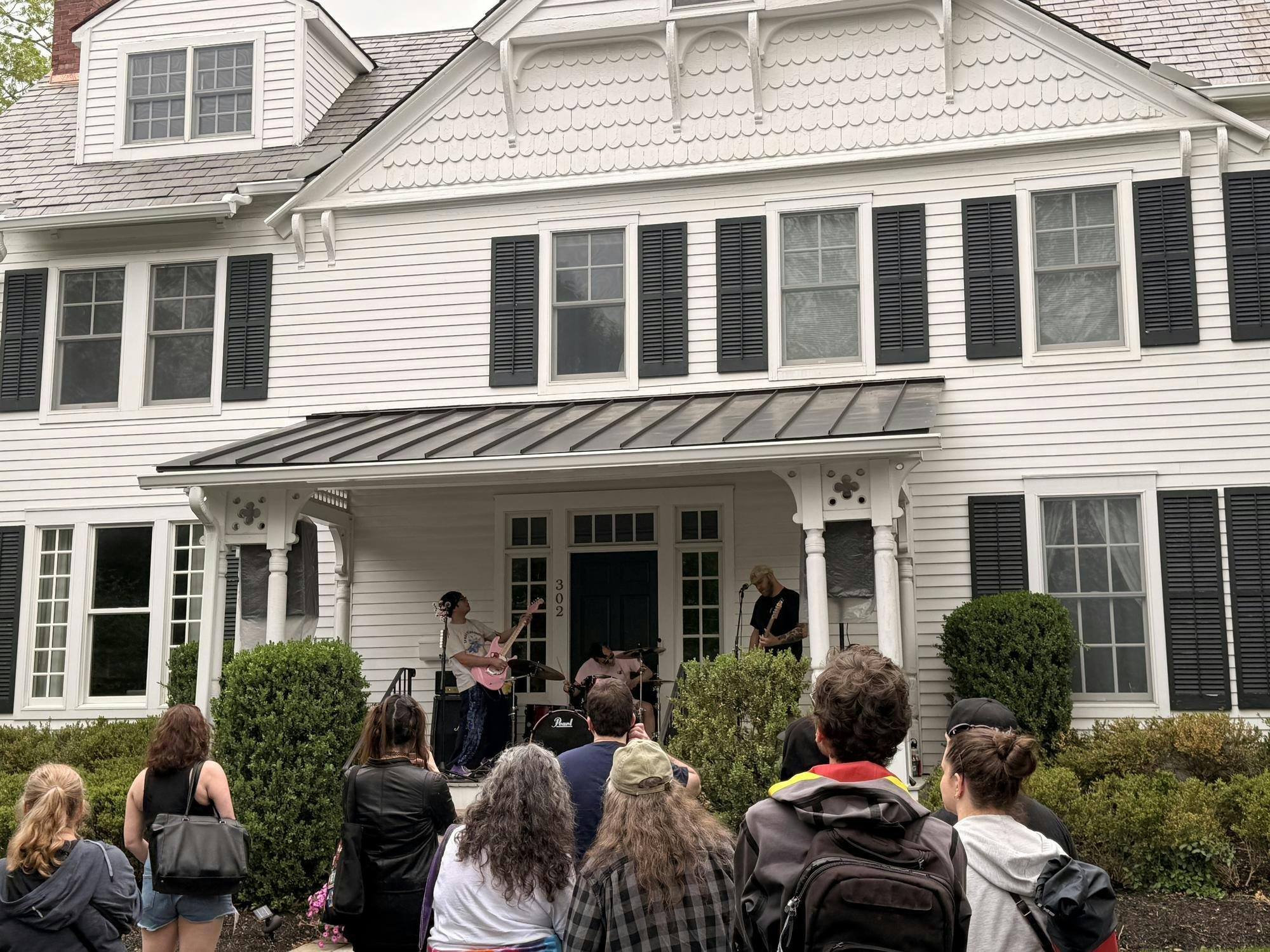 A crowd of spectators are gathered in front of a large white house where three musicians playing the guitar and drums are standing and performing on the porch.