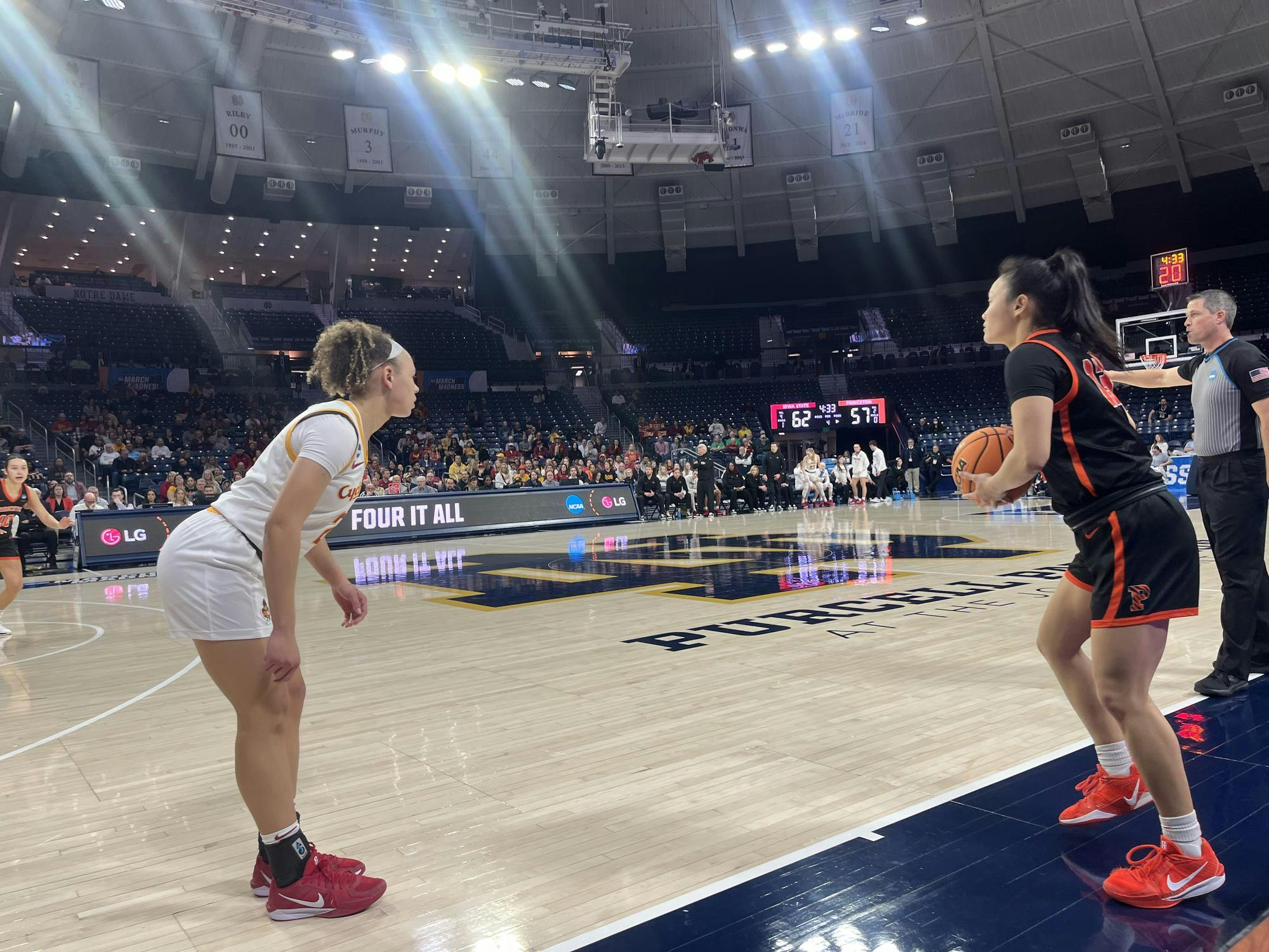 Basketball stadium with female basketball player wearing black and orange holding the ball on the right side and another female basketball player wearing white in a low defensive stance on the left side.
