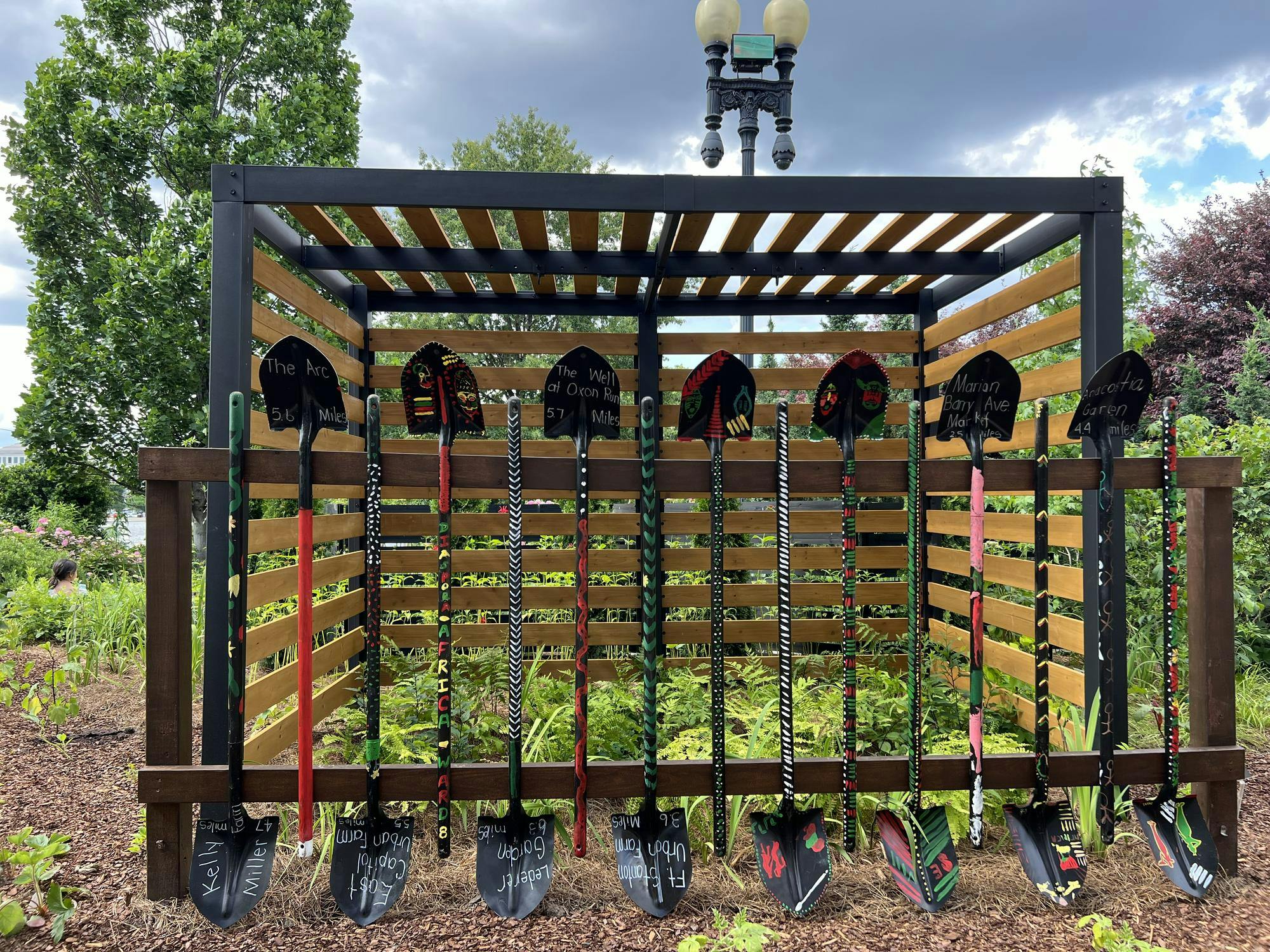 A collection of decorated shovels stand before a wooden enclosure with plants in the background at the Botanical Gardens in Washington, DC.