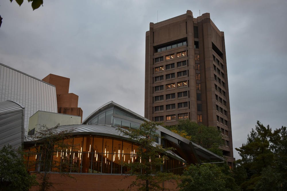 In the foreground, a glass-covered building with vertical bar lights inside. On the right, a brutalist-style gray building with narrow, horizontal black windows. In the background, a cloudy sky looms.