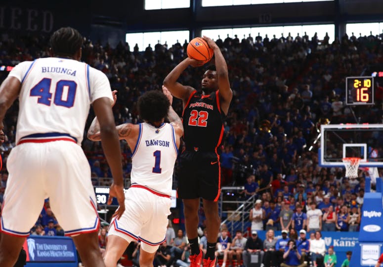 A player in a black jersey jumping and shooting a basketball in front of a player in a white jersey.