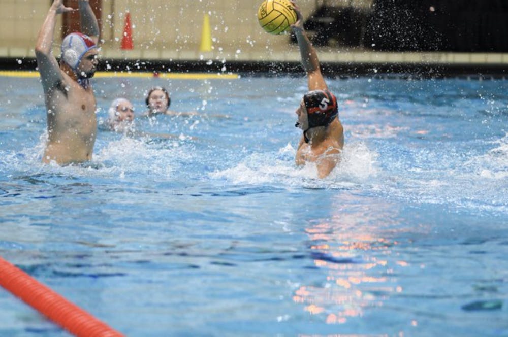 Two players are in the foreground of the pool. A Tigers player, holding the ball above his head, tries to get the ball past opposing team defender. Opposing team defender has both hands outstretched to block the ball. A Princeton Tigers player and LIU player are visible in the background facing the ball.
