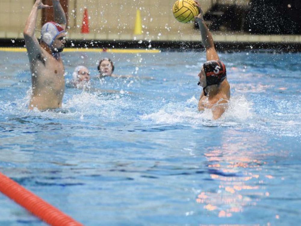 Two players are in the foreground of the pool. A Tigers player, holding the ball above his head, tries to get the ball past opposing team defender. Opposing team defender has both hands outstretched to block the ball. A Princeton Tigers player and LIU player are visible in the background facing the ball.