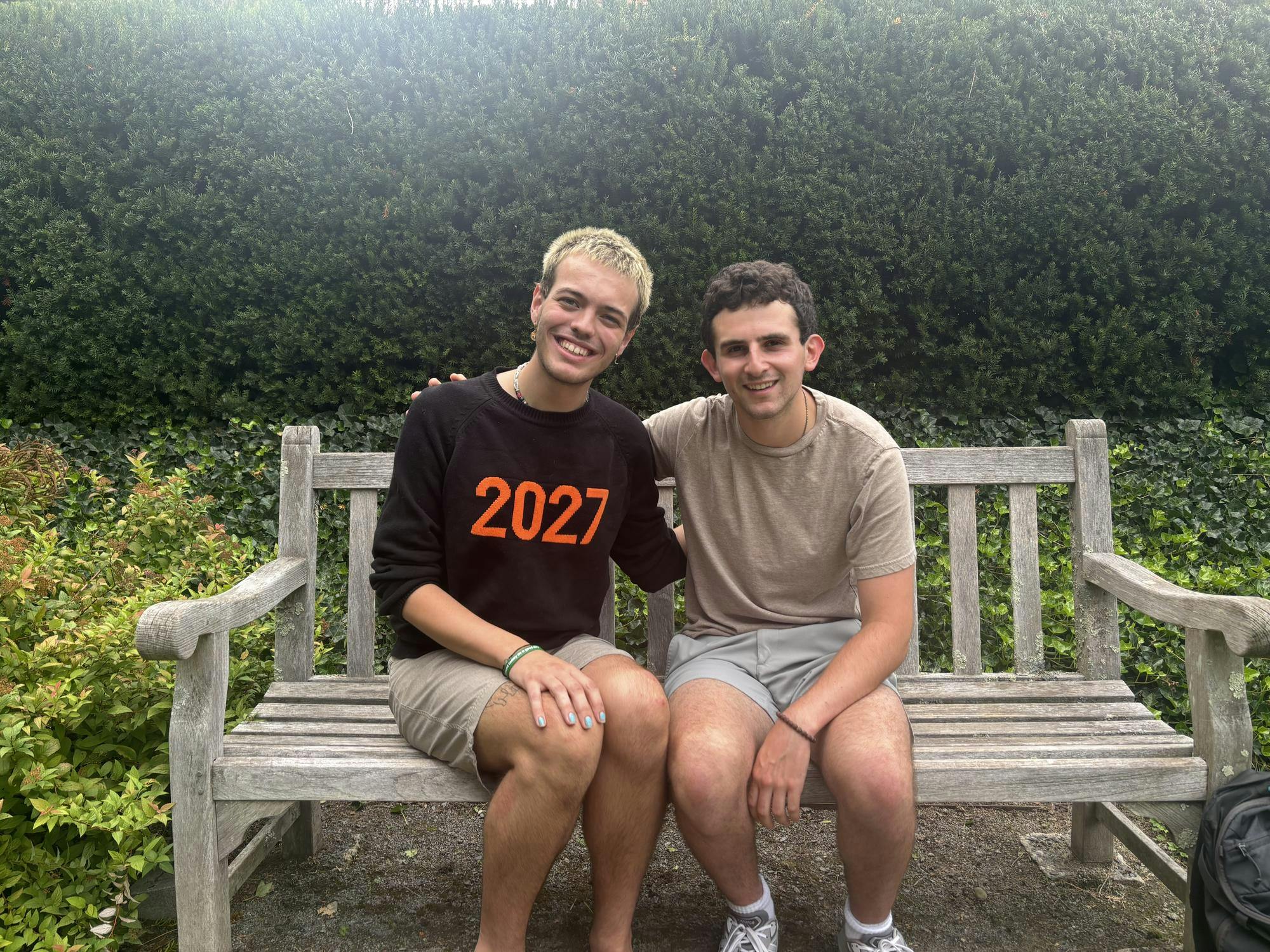 Two men sitting on a bench with green trees in the background.