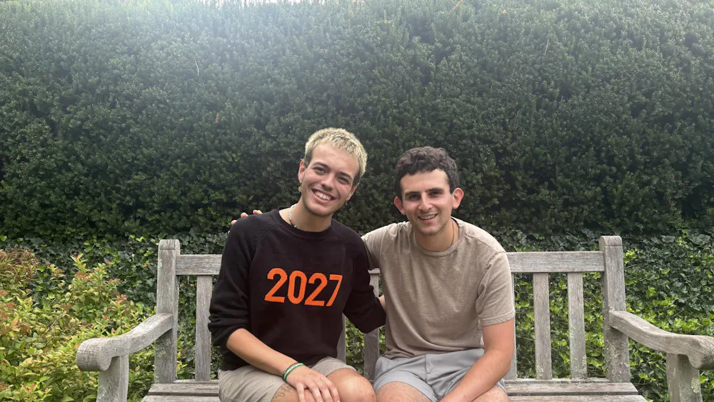 Two men sitting on a bench with green trees in the background.
