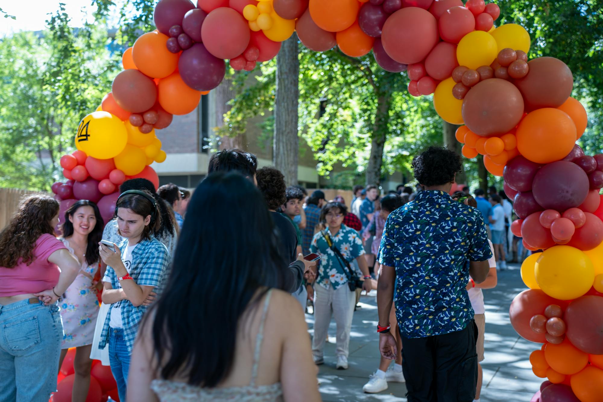 Students walk under red, orange, and yellow balloon arch, on a tree-lined path.