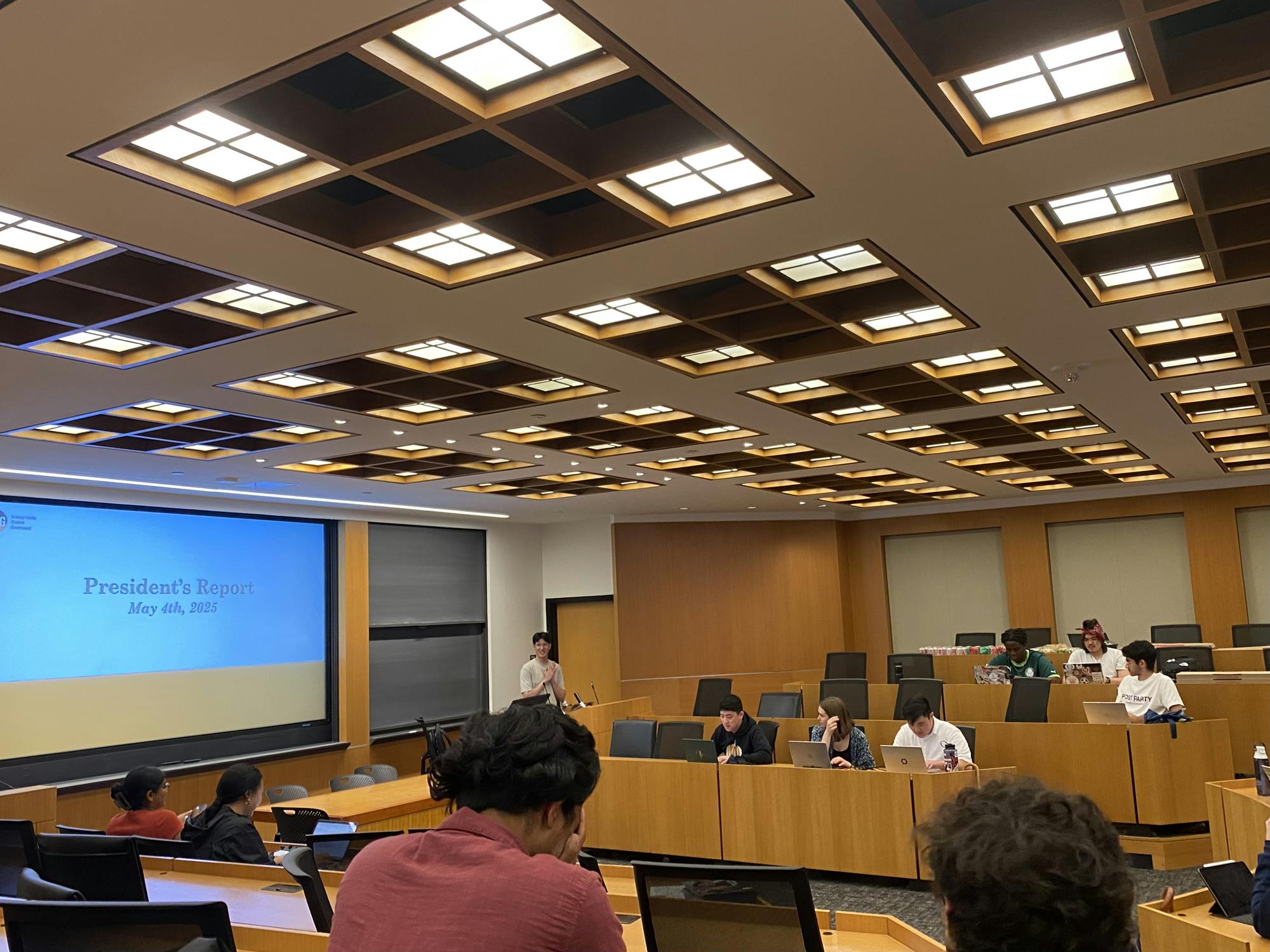 A screen reads “President's report. May 4, 2025.” People sit at wooden desks with their computers open.