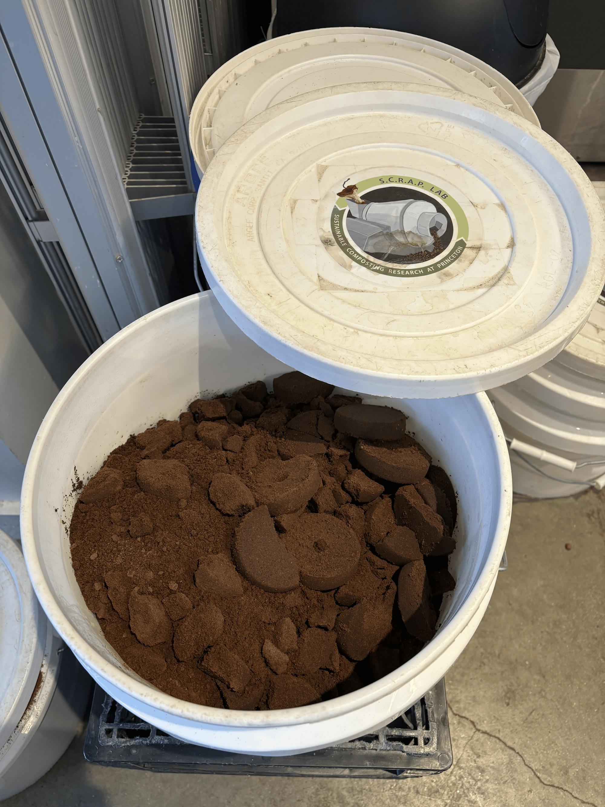 A white bucket full of coffee puck remains with a lid partially over the opening and several more buckets in the background.